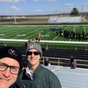 2 men in front of football field with marching band on it